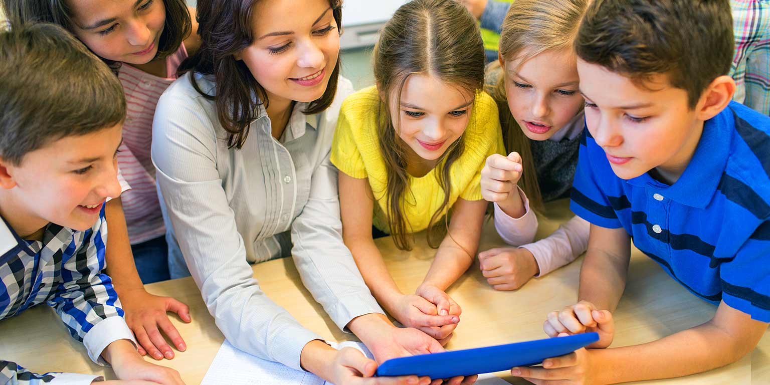 A teacher and a group of children look at a tablet computer in a classroom. 